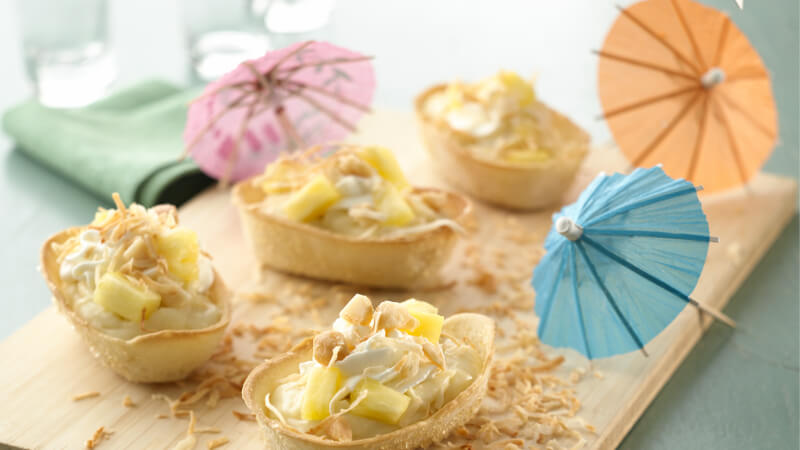 Mini Old El Paso Taco Bowls willed with coconut cream, pineapple chunks and shredded toasted coconut on a wooden serving board and mini colourful umbrellas
